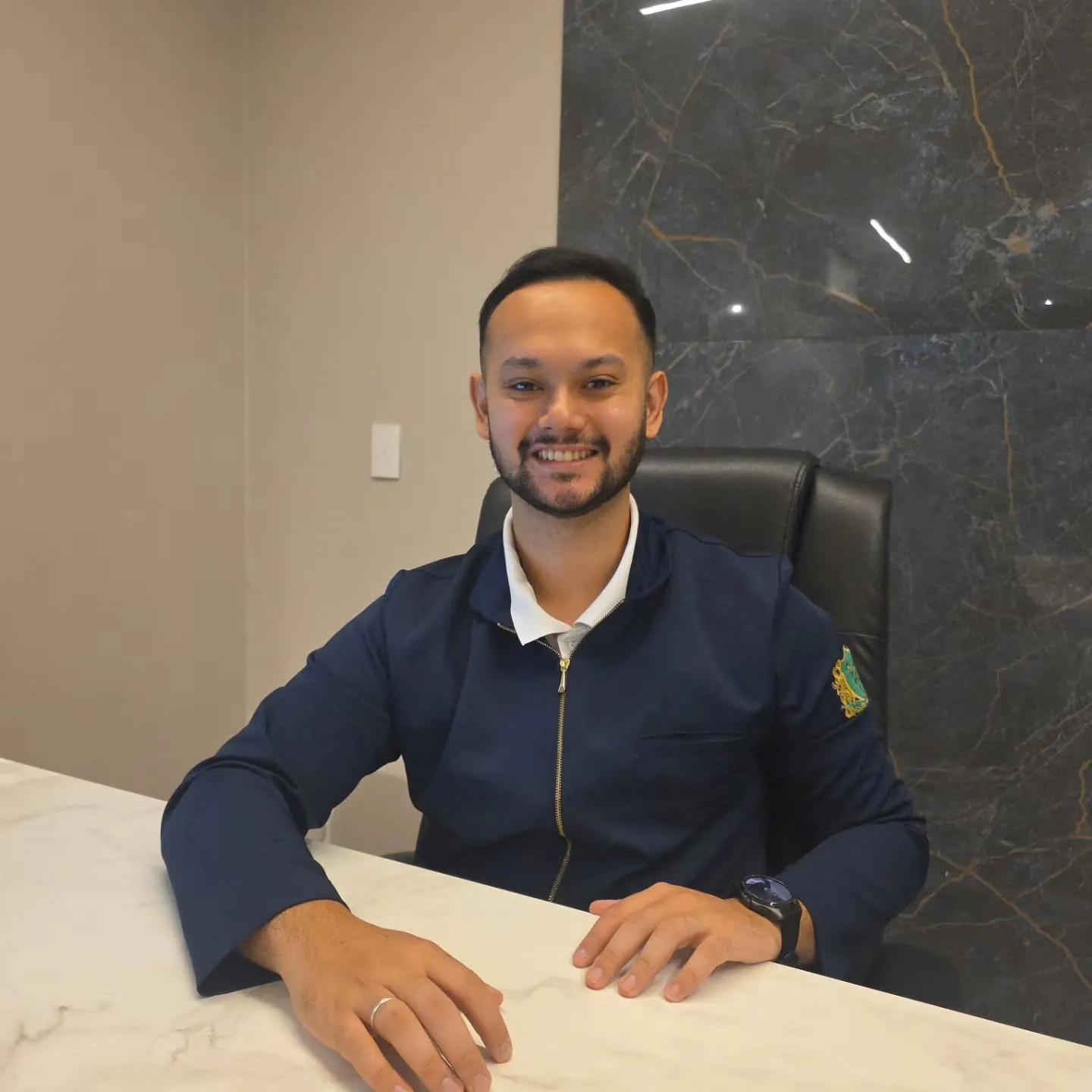 Dr. Giulian Saito, fisioterapeuta, sorrindo sentado à mesa do consultório